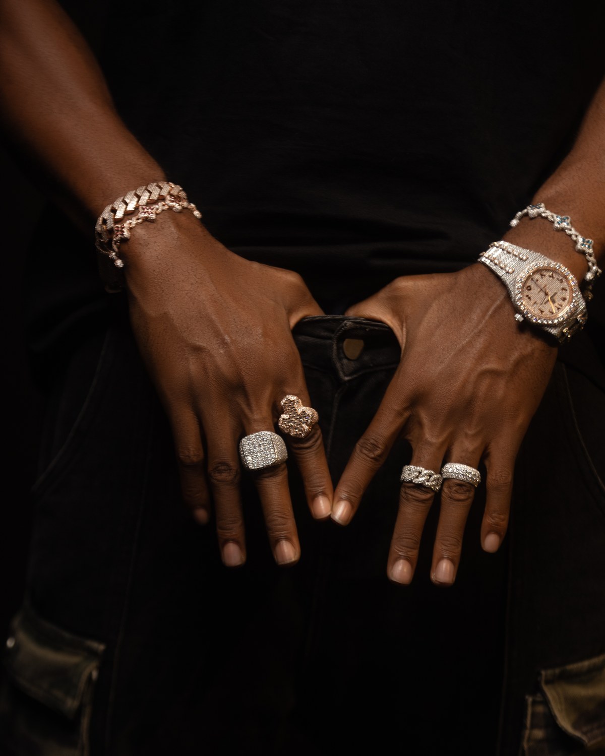 Hands wearing bracelets, rings, and a watch against black.